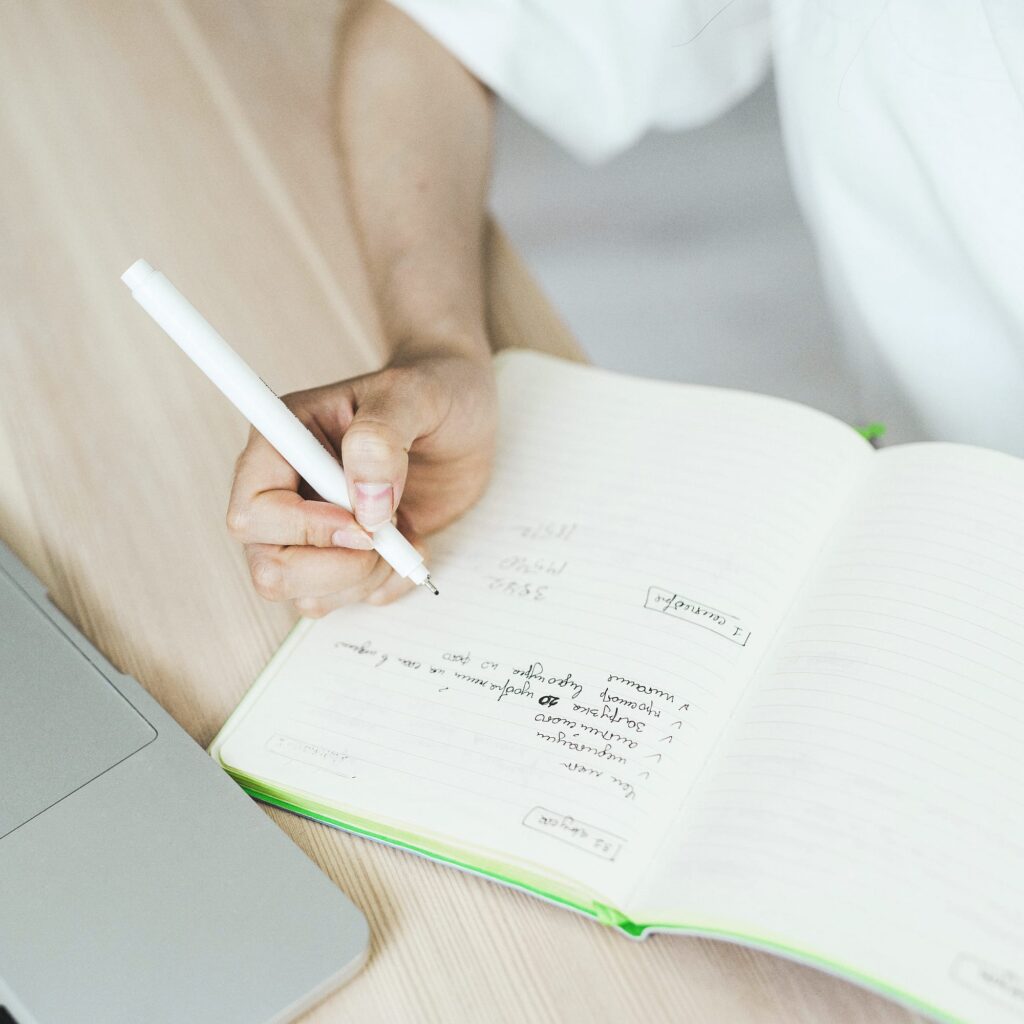 A hand writes a list in a notebook next to a laptop on a light wooden desk.