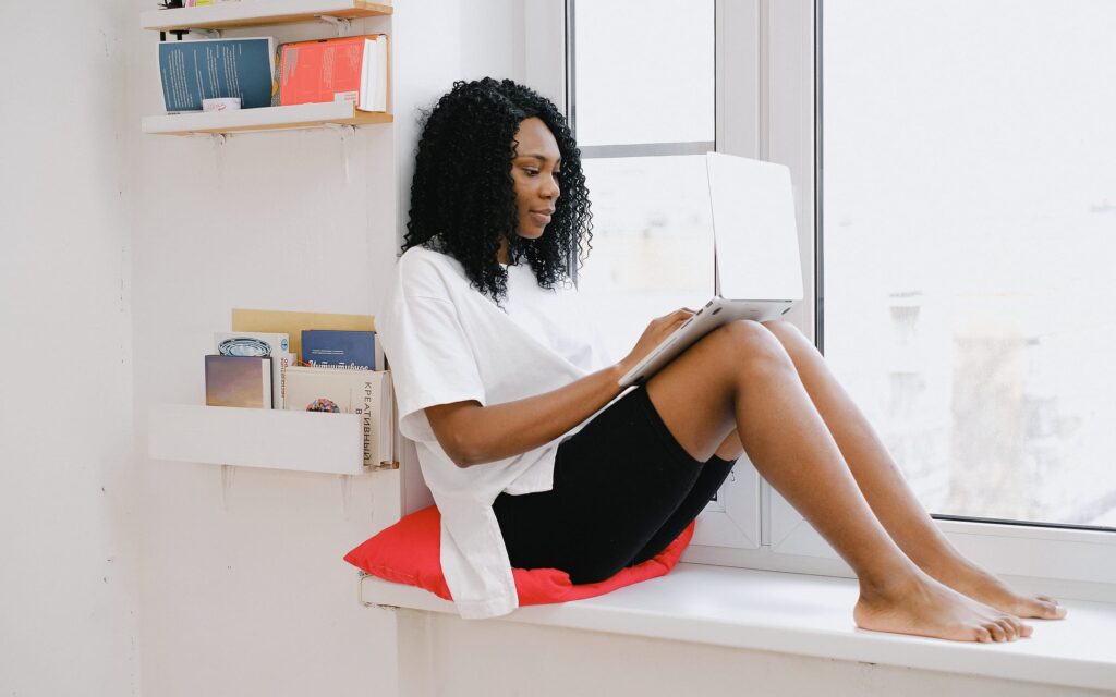 A woman sits on a bright window ledge, reading on a laptop.