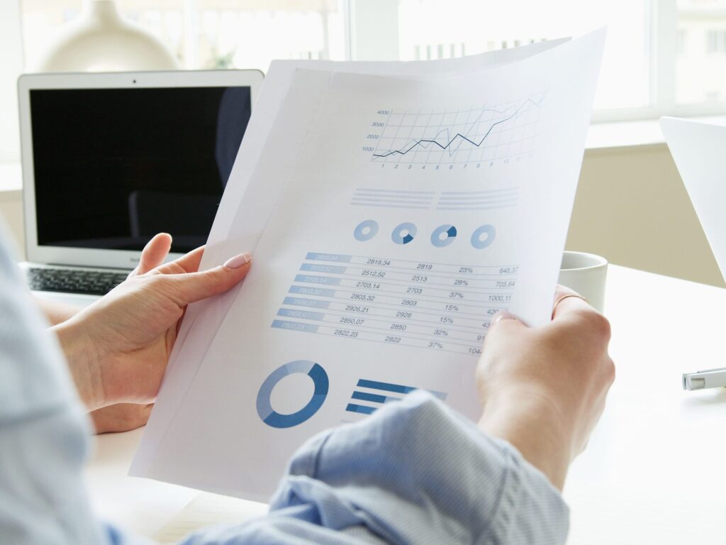 Hands hold a printed report with line, donut and bar charts, a laptop in the background.